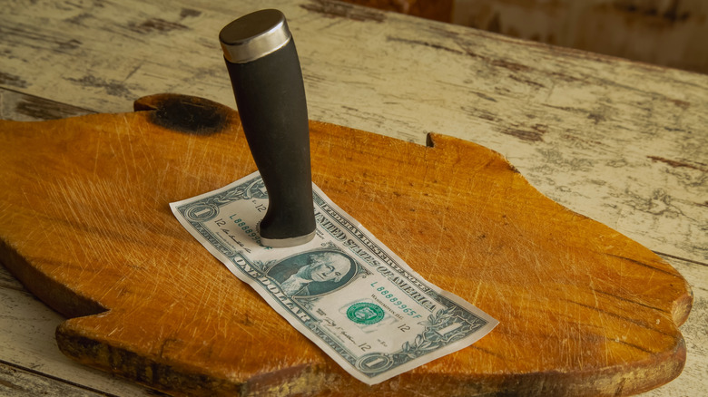 A broken kitchen knife pins a dollar bill to a cutting board