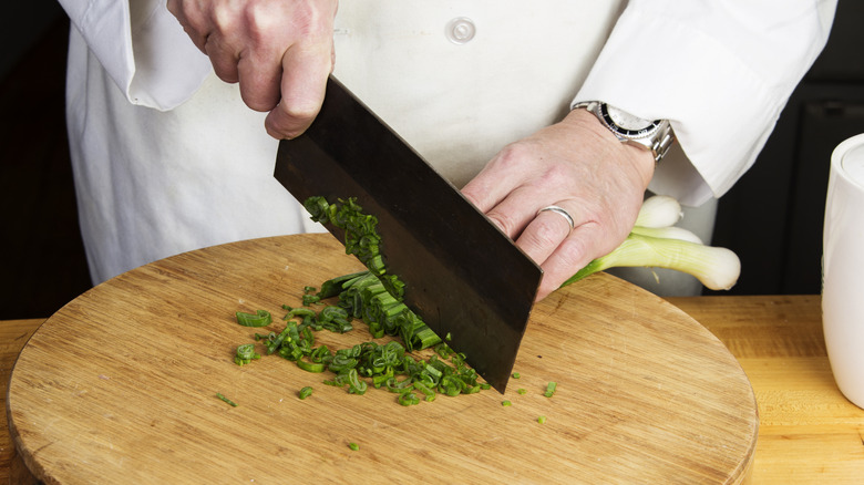 A chef's hands use a Chinese cleaver to chop scallions