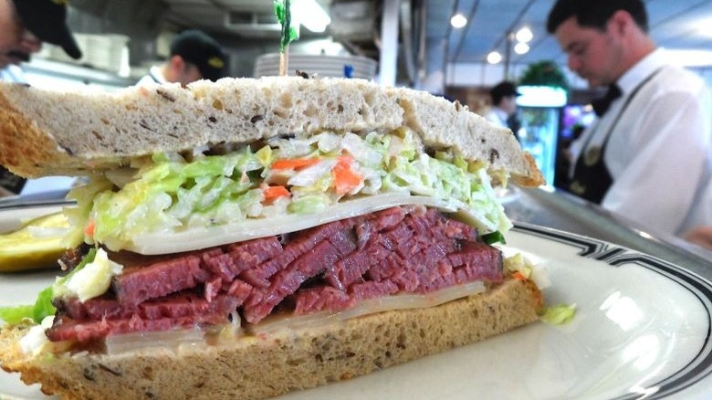 Pastrami sandwich with cheese and coleslaw on a plate, with a workers in the background